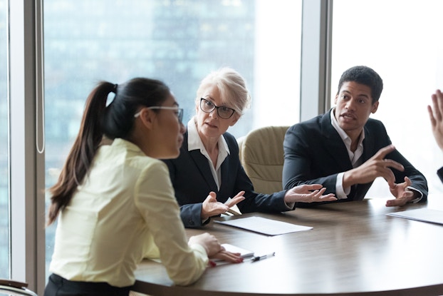 A trio of work colleagues sits around a conference room table. The two facing the camera -- a woman in glasses and a young man -- both look irate. They are gesturing and speaking with impatient and displeased looks on their faces. On the far left, facing them, is a young woman with her hair in a ponytail and her hands folded on the table.