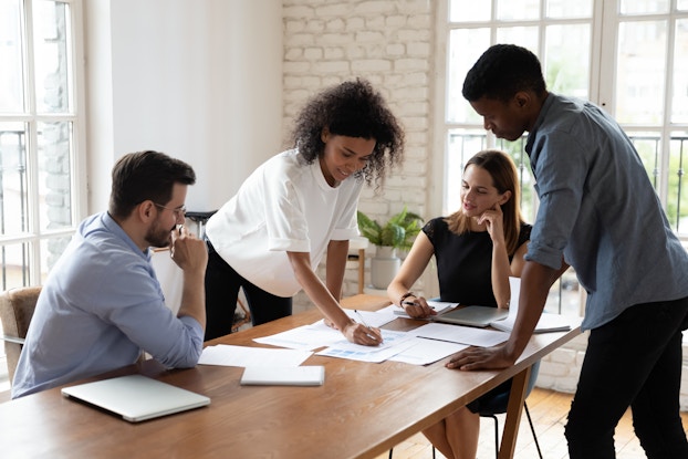 Four coworkers stand or sit around a table spread with various papers. One of the people standing, a woman in a white blouse, writes something on one of the papers as her colleagues look on.