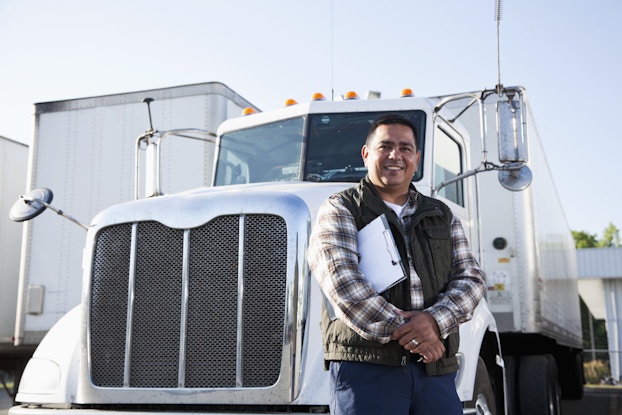 A man with a clipboard under one arm stands smiling in front of the cab of an 18-wheel truck.